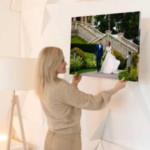 A woman hangs a framed wedding photo on a white wall. Printed as a Single ChromaLuxe Indoor Metal Photo Print, it shows a bride and groom walking near outdoor stairs and greenery. Choose from any base, size, and finish for your metal prints. Prints 4 You