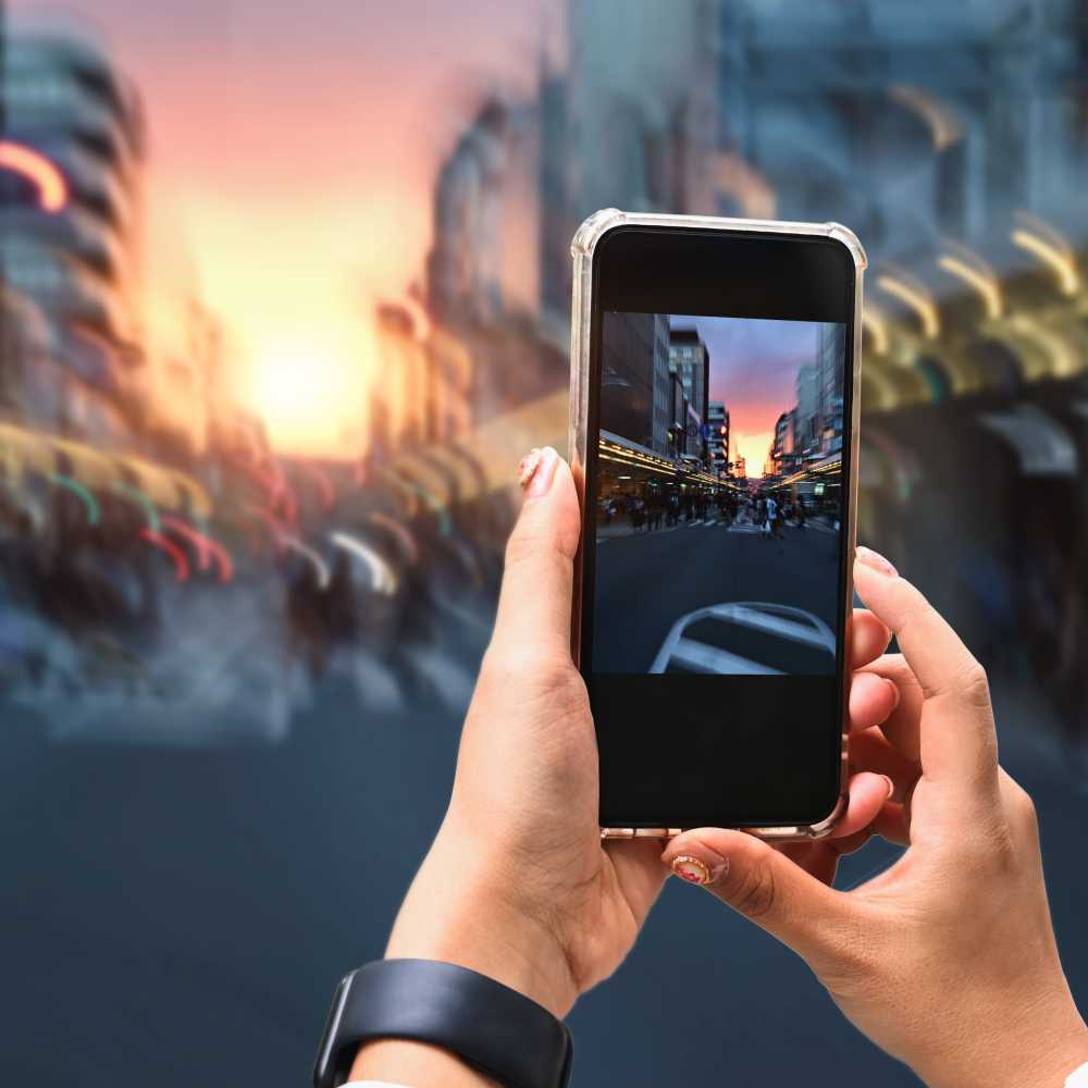 A person holding a smartphone takes a photo of a city street at sunset, with blurred buildings and lights in the background—perfect for turning into stunning Metal Photo Prints, made by Prints 4 You. Prints 4 You