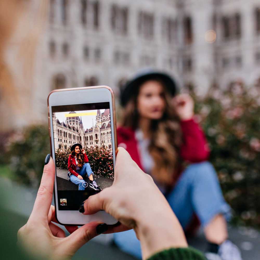 A person holds a smartphone, taking a photo of a woman in casual clothes and a hat, sitting outdoors by flowers and a blurred historic building. Capture moments like this forever with Metal Photo Prints made by Prints 4 You.