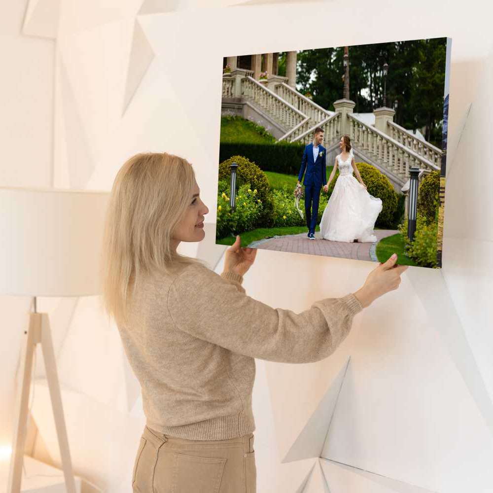 A woman hangs a Chromaluxe Metal Print on a white wall. The framed photo captures a bride and groom walking hand-in-hand outdoors, dressed in formal wedding attire. Prints 4 You