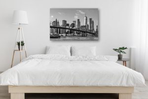 A modern, minimalistic bedroom features a large bed with white bedding, a floor lamp, side table with books, a potted plant, and striking photos on metal—a black-and-white cityscape artwork above the bed completes the look. Prints 4 You