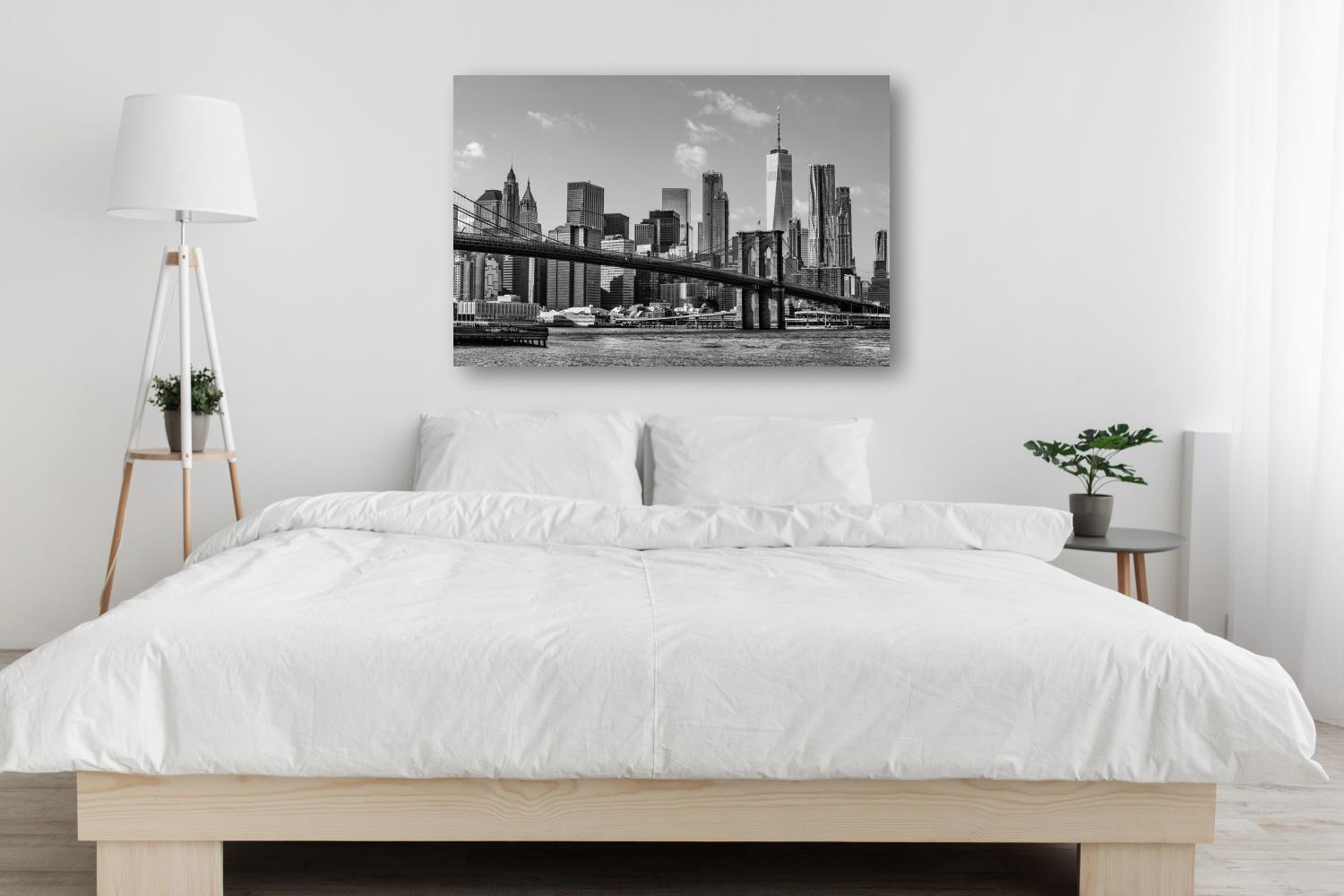 A modern, minimalistic bedroom features a large bed with white bedding, a floor lamp, side table with books, a potted plant, and striking photos on metal—a black-and-white cityscape artwork above the bed completes the look. Prints 4 You