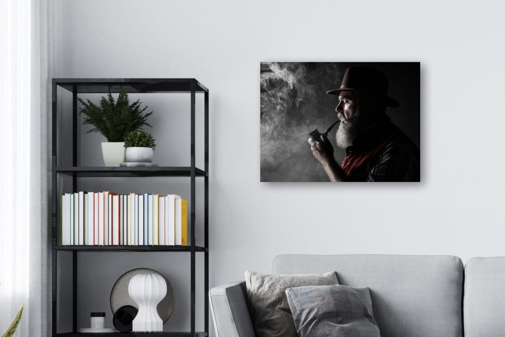 A modern living room with a gray sofa, a black shelving unit with books and plants, and a stunning Chromaluxe Metal Print of an older man with a beard wearing a hat and smoking a pipe mounted on the wall. Prints 4 You