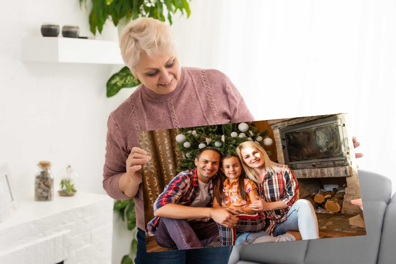 An older woman smiles as she holds a framed family photo of a man, woman, and child sitting together by a decorated Christmas tree and fireplace. Order custom photo prints to cherish your own special memories. Prints 4 You