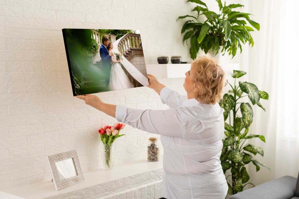 An older woman hangs a framed wedding photo on a bright living room wall among plants and family pictures—each beautifully preserved with Single ChromaLuxe Indoor Metal Photo Prints, available in any base, size, or finish. Prints 4 You
