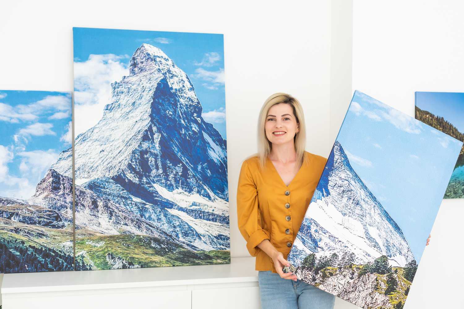 A woman in a yellow top smiles while holding a canvas print of a snowy mountain landscape, standing beside larger Metal Photo Prints of mountain scenes displayed on a white wall. Prints 4 You