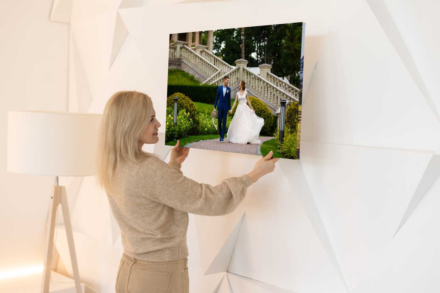 A woman hangs a Chromaluxe Metal Print of a bride and groom walking outdoors on a white, geometric-patterned wall. A floor lamp sits nearby, highlighting the stunning photo on metal. Prints 4 You