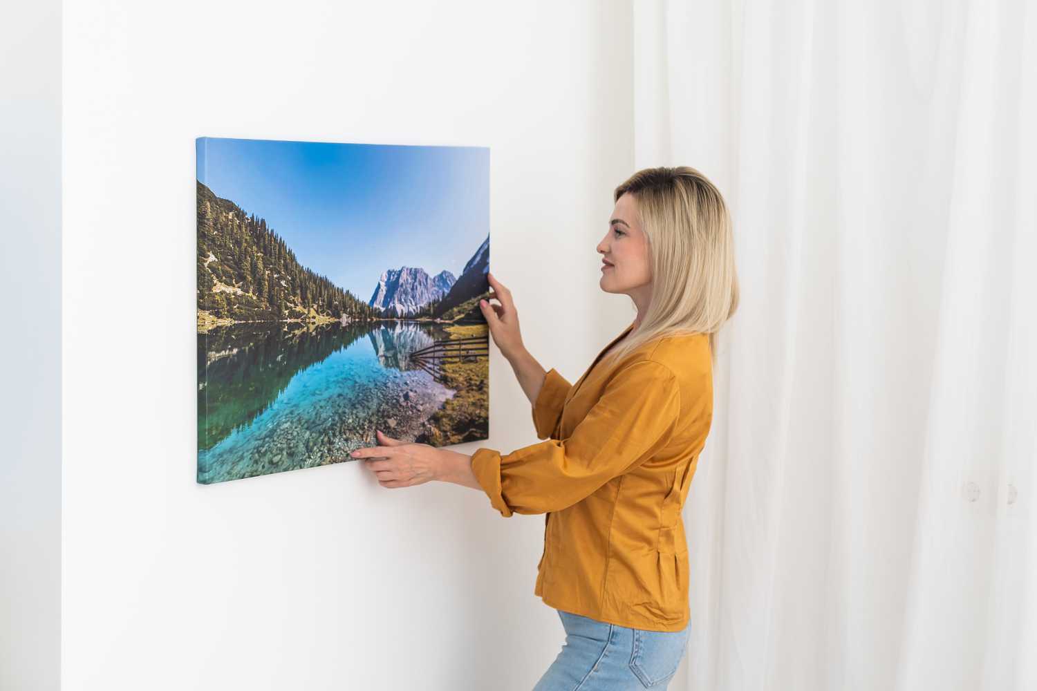 A woman in a mustard yellow shirt hangs a framed landscape photo of a lake and mountains on a white wall, showcasing the vibrant detail possible when you order custom photo prints. Prints 4 You