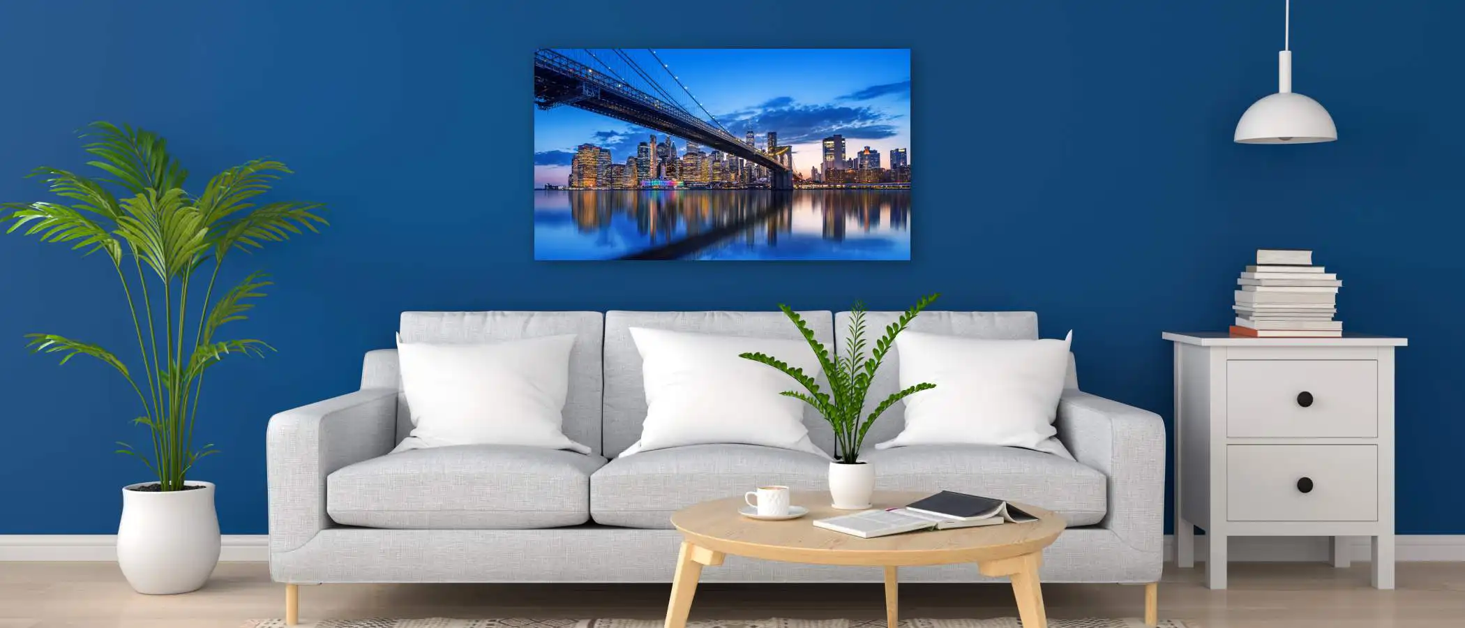 A modern living room with a light gray sofa, white pillows, a round wooden coffee table with potted plants, and a cityscape photo on a blue wall. A white lamp hangs by a small bookshelf on the right. A metal photo print made by Prints 4 You
